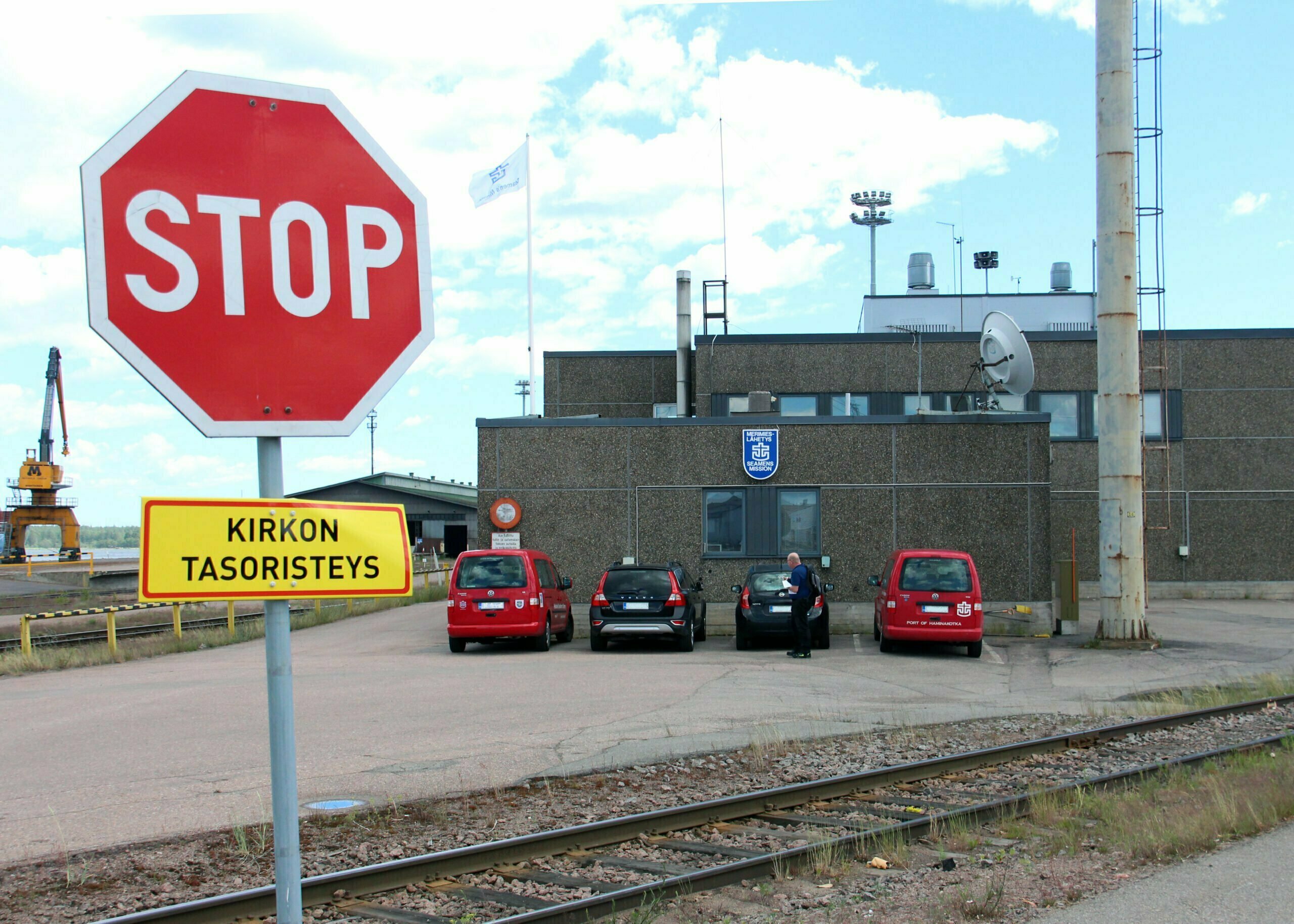 Ruskea betonielemenntirakennus, jonka vieressä neljä autoa ja kuvan etualla junaraide ja liikennemerkki Stop, jossa lisäkyltti Kirkon tasoristeys.
