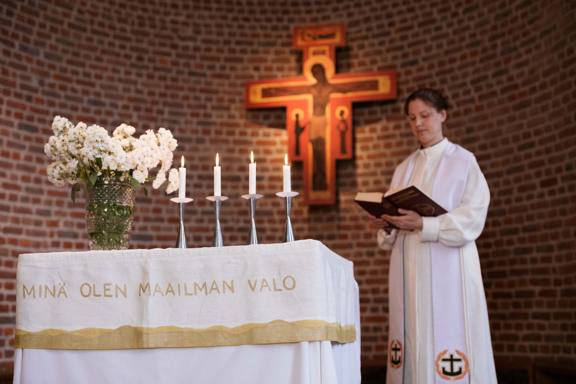 "Pappi sekä alttaripöytä, jossa valkoinen liina, valkosia kukkia ja neljä kynttilää taustalla punatiiliseinällä risti."