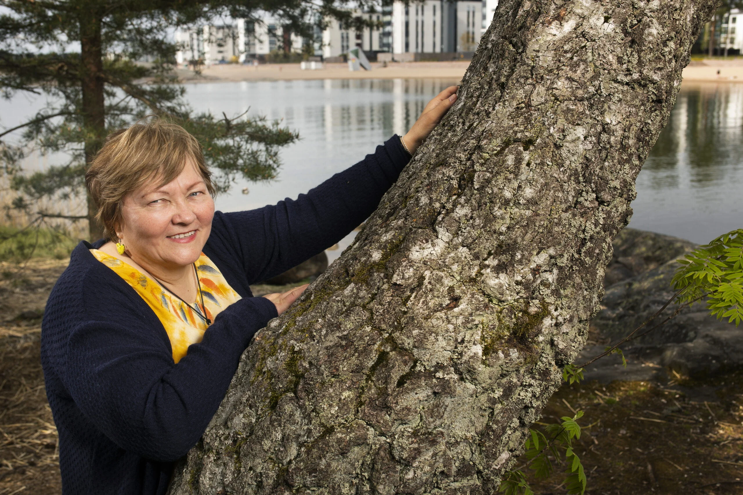 Hymyilevä Pirjo-Sisko Katjaisto nojailee männyn runkoon Hensingissä. Taustalla on Aurinkolahden rantaa ja kerrostaloja.