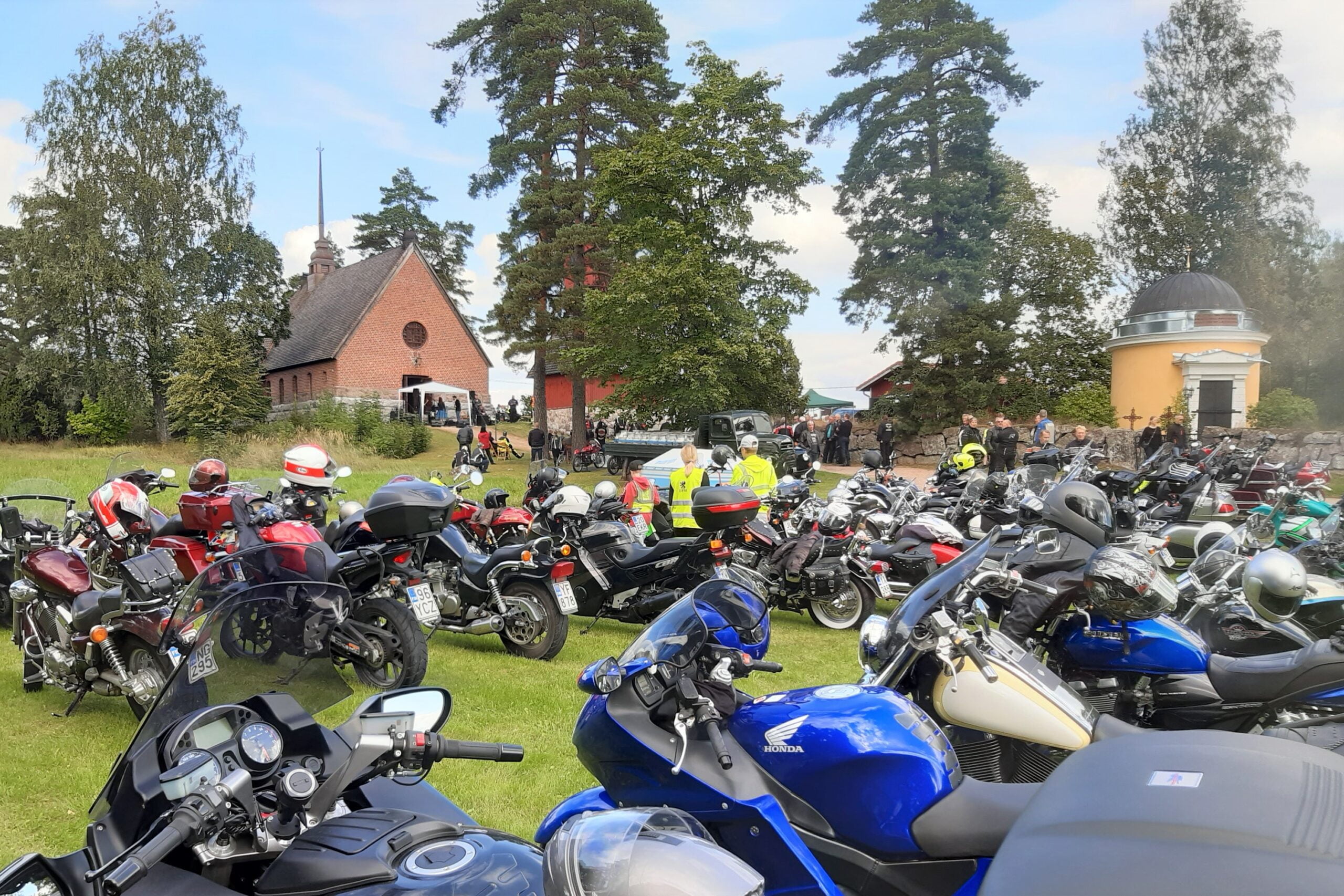 "Kirkon edessä suuri määrä erivärisiä moottoripyöriä ja kirkkoon meneviä motoristeja."