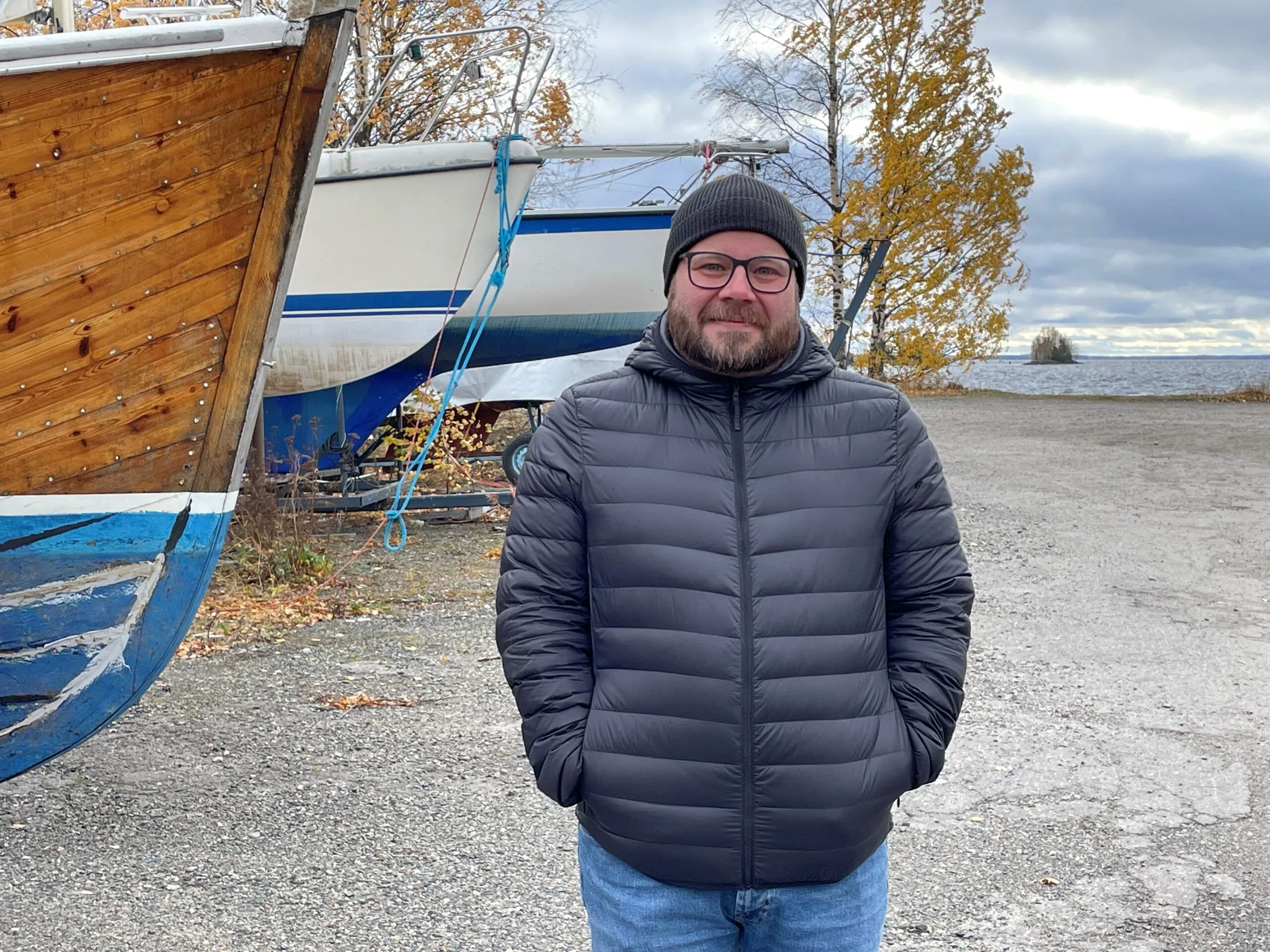 "Parrakas silmälasipäinen mies yllään kevytuntuvatakki ja pipo katsoo kameraan taustalla talviteloille nostettuja veneitä ja syksyisiä koivuja sekä järvi".