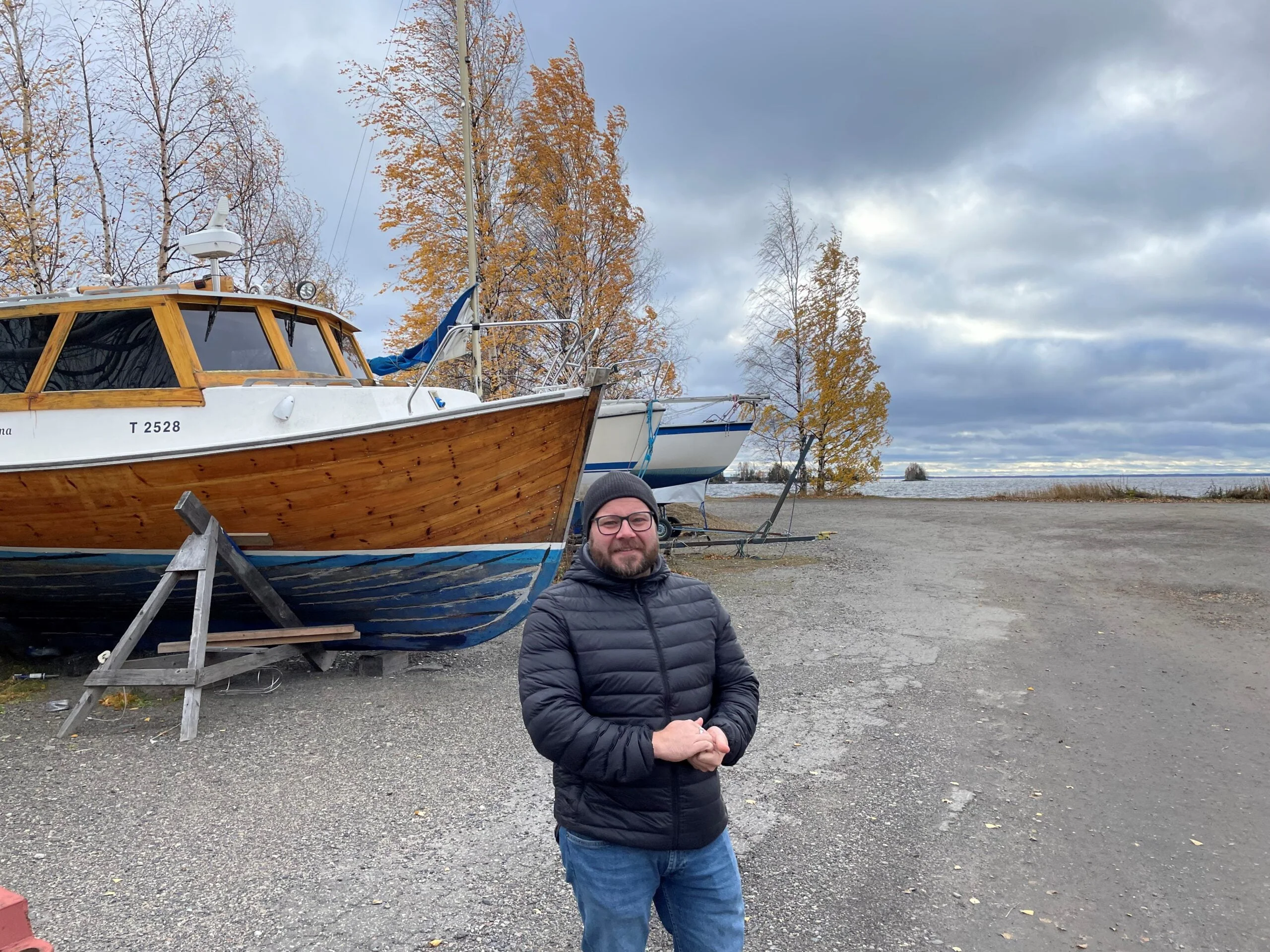 "Parrakas silmälasipäinen mies yllään kevytuntuvatakki ja pipo katsoo kameraan taustalla talviteloille nostettuja veneitä ja syksyisiä koivuja sekä järvi".