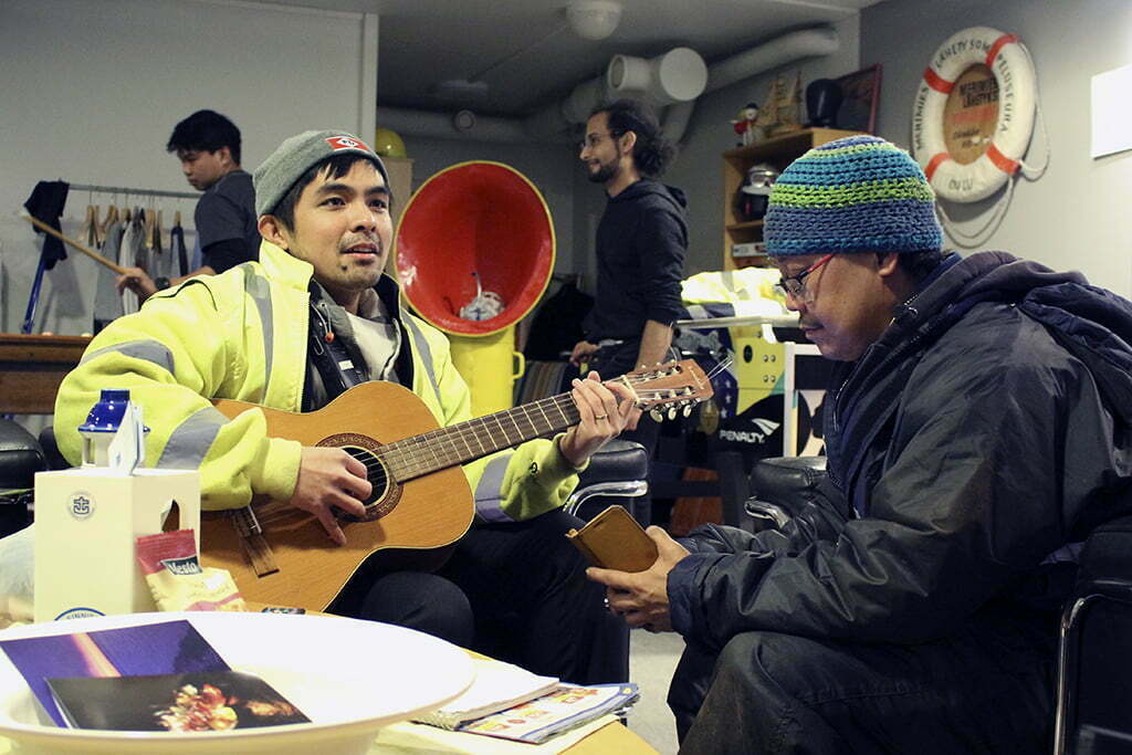 "Etualla kaksi aasialaista miestä, joista toinen soittaa kitaraa ja toinen katsoo kännykkää. Taustalla kaksi mieshenkilöä pelaa biljardia."