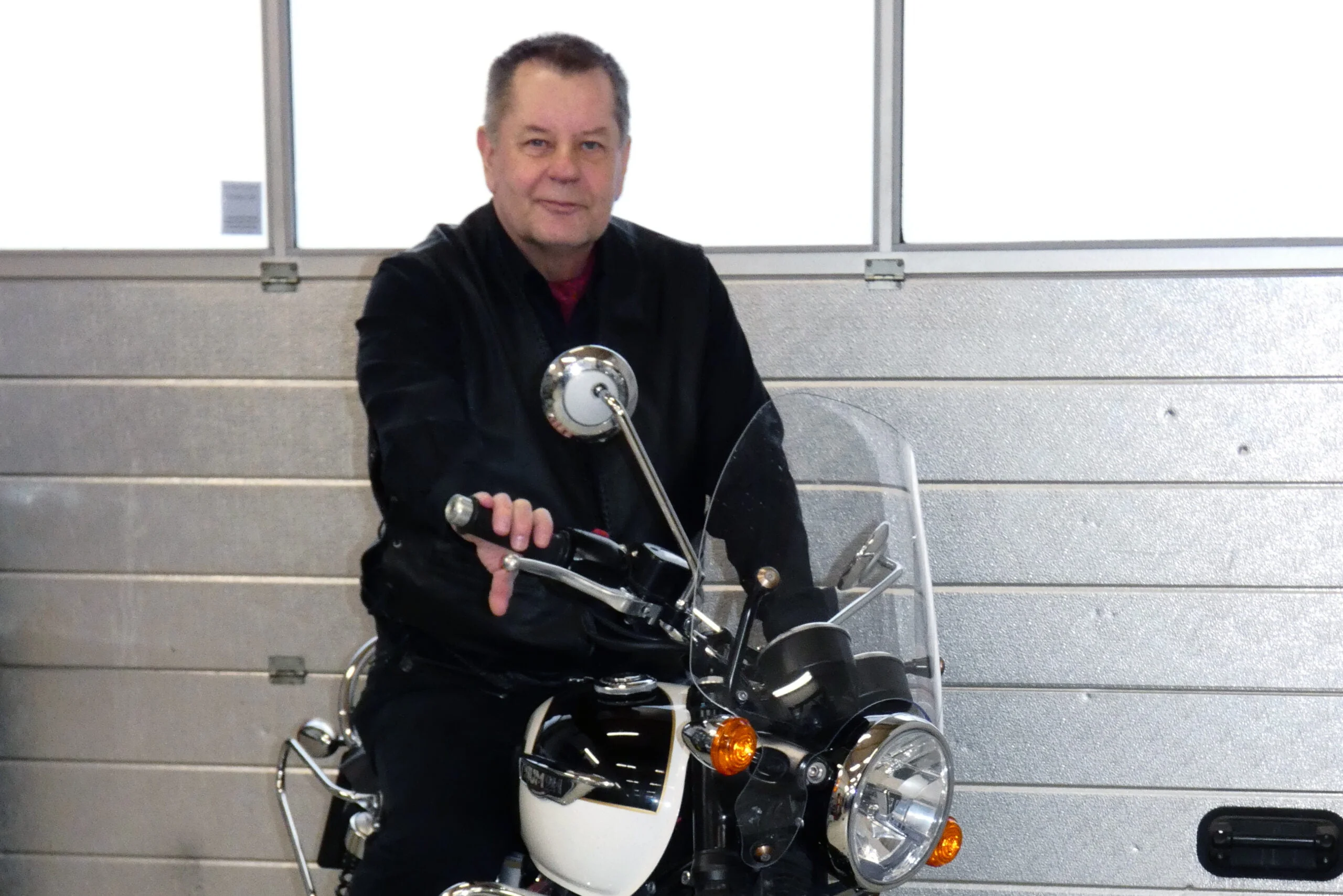 "Mies istuu valkoisen moottoripyörän päällä pitäen toisella kädellä kiinni ratista. Taustalla metallinen ovi".
