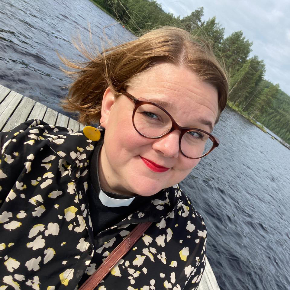 "Hymyilevä silmälasipäinen keski-ikäinen nainen, jolla on papin panta kauluksessa, seisoo järven rannalla laiturilla".