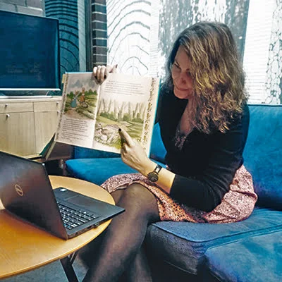 "Ruskeatukkainen nainen istuu sinisellä sohvalla avoin kannettava edessään tietokone, jonne nainen näyttää satukirjaa ja osoittaa sormellaan sivun sienikuvaa".