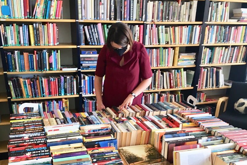 "Kasvomaski yllä oleva punapaitainen nainen tutkii pöydällä olevia kirjoja. takanaan seinällä olevassa kirjahyllyssä on runsaasti kirjoja."