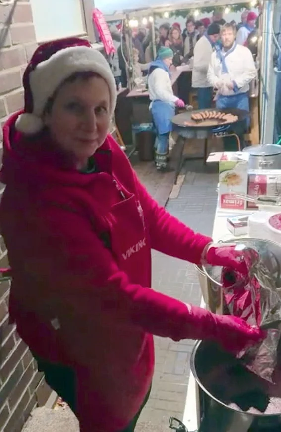 "Punaisiin jouluvaatteisiin sonnustautunut hymyilevä nainen valmistamassa glögiä pihakojussa."