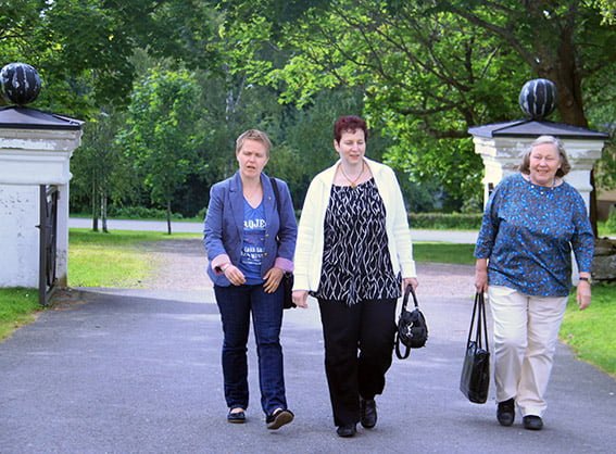 "Kolme naista kävelemässä kirkon kiviportista sisään. Taustalla vihreitä lehtipuita."