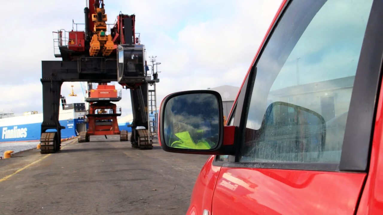 Punainen auto, jonka sivupeilistä näkyy kuljettaja, ajaa laiturialueella kohti laivaa.