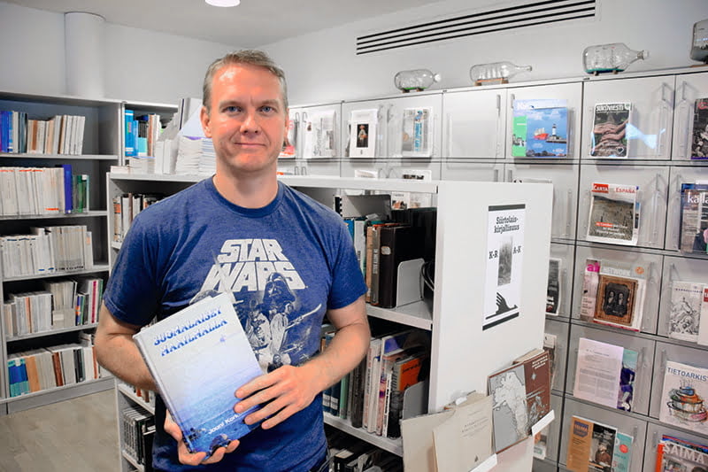 "Siniseen Star wars -paitaan pukeutunut hymyilevä mies pitää käsissään kirjaa Suomalaiset maailmalla. Taustalla valkoisissa hyllyissä kirjoja ja lehtiä."