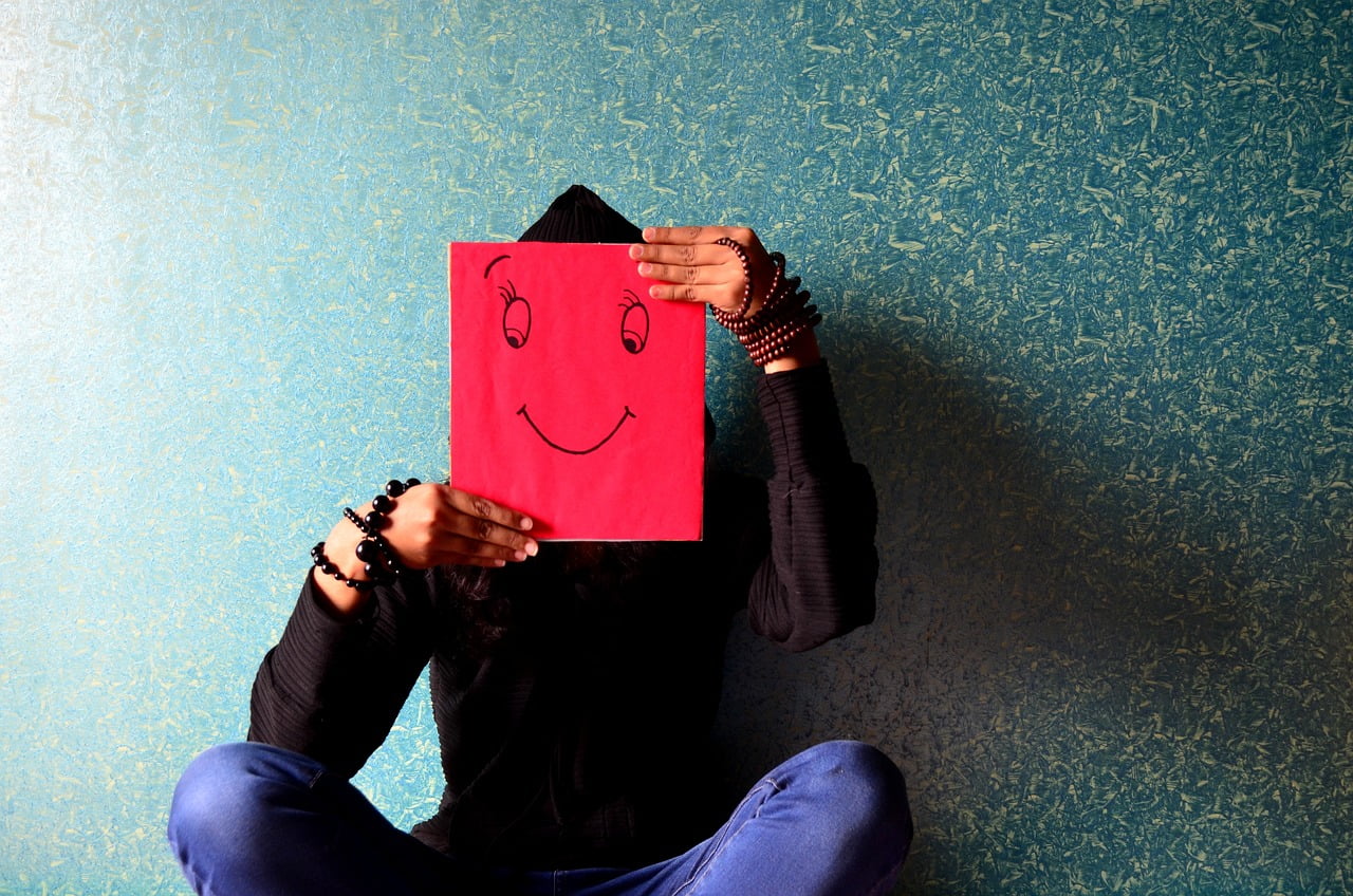 Seinää vasten istuva mies pitelee kasvojensa edessä punaista paperia, johon piirretty hymyemoji.