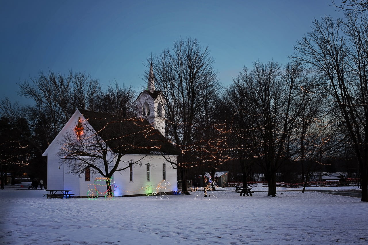 Valkoinen kirkko jouluvaloineen hämärässä lumisessa maisemassa.