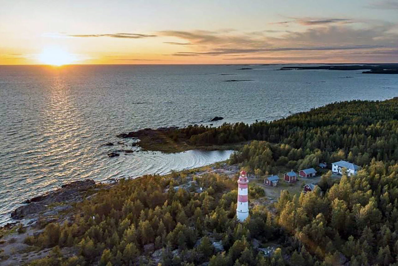 Ilmasta otettu kuva, jossa aurinko laskee meren horisonttiin ja etualla maassa on majakka ja muutama pieni rakennus.