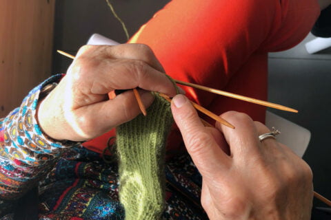 Lähikuva seniorin käsistä, jotka neulovat vihreää villasukkaa.