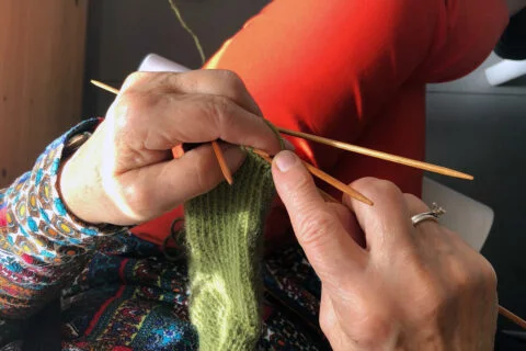 Lähikuva seniorin käsistä, jotka neulovat vihreää villasukkaa.