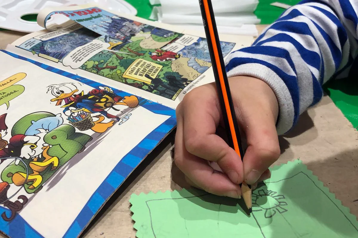 Lähikuva lapsen kädestä, joka piirtää vihreälle paperille vieressään pöydällä Aku Ankka -lehti.