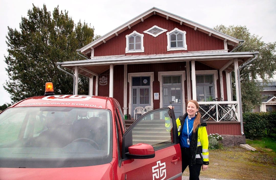 Punaisen kaksikerroksisen puutalon edessä punainen auto, jonka etuovea pitää auki hymyilevä nainen.