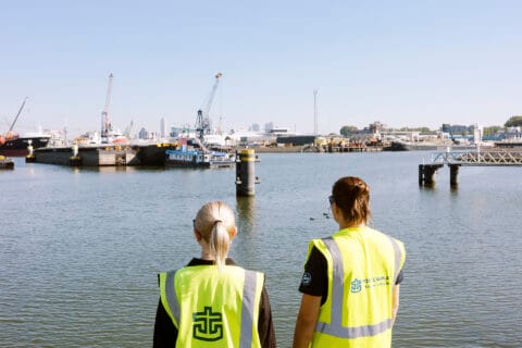 Kaksi keltaiseen Port Chaplain -tekstilliseen huomioliiviin pukeutunutta naista katselee satama-altaan toisella puolella olevia laivoja.