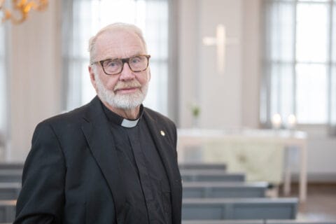 Harmaapartainen mies pukeutuneena mustaan papin asuun, taustalla kaksi ikkunaa, joiden välissä seinällä risti.