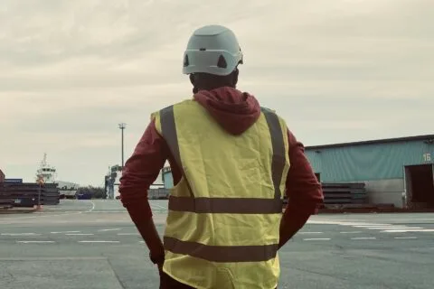 Mies valkoisessa kypärässä ja keltaisessa huomioliivissä satama-alueella, takaa päin otettu kuva.