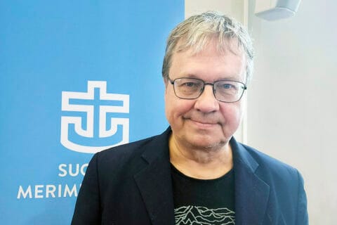 Vaaleahiuksinen, silmälasipäinen, hymyilevä mies sinisen roll upin edessä, jossa Merimieskirkon ankkuriristi.