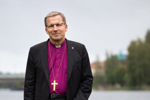 Kuvassa seisoo pappi, jolla on päällään musta puku ja violetti kaulus. Hänellä on ristiriipus kaulassaan ja taustalla näkyy järvi ja puustoa.