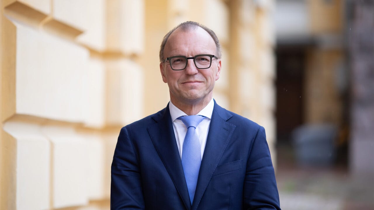 Silmälasipäinen, siniseen puvuntakkiin ja vaaleansiniseen solmioon pukeutunut mies.