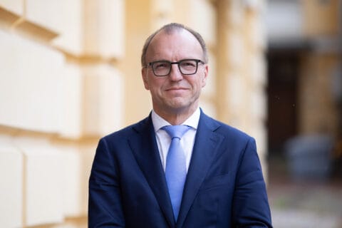 Silmälasipäinen, siniseen puvuntakkiin ja vaaleansiniseen solmioon pukeutunut mies.