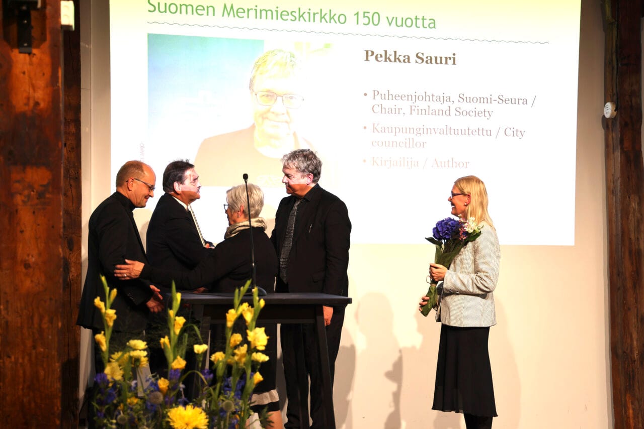 Esiintymislavalla viisi henkilöä, joista yhdellä kukkapuska sylissään.