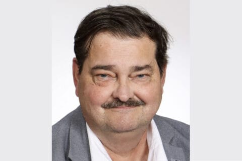 Tummatukkainen, harmaaseen pikkutakkiin ja valkoiseen paitaan pukeutunut hymyilevä mies.