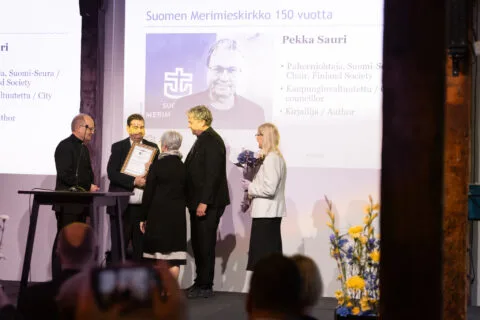 Kaksi henkilöä ottaa vastaan kehystetyn diplomin kolmelta henkilöltä, joista yhdellä on kukkapuska kädessään.