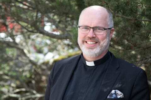 Mustaan takkiin ja mustaan papinpaitaan pukeutunut harmaapartainen ja -tukkainen mies taustallaan vihreitä puunoksia.