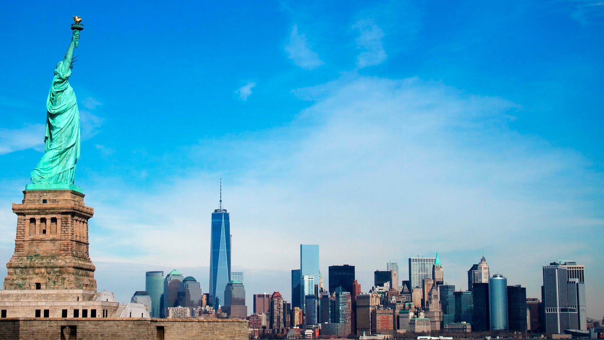 Vasemmalla vihertävä iso vapaudenpatsas-monumentti ja taustalla kaupungin silhuetti, sininen taivas.
