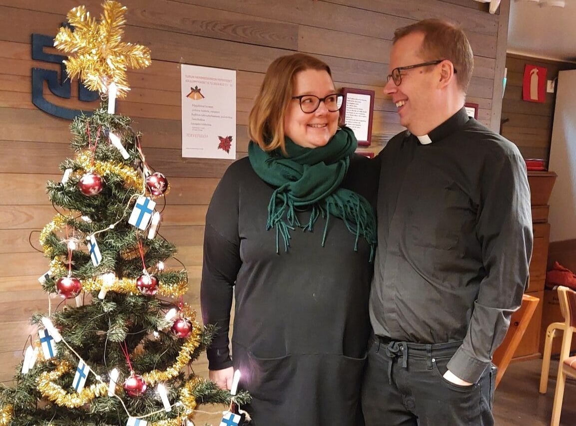 Toisiinsa katsovat, hymyilevät mustiin papinpaitoihin pukeutuneet nainen ja mies koristellun joulukuusen vieressä.