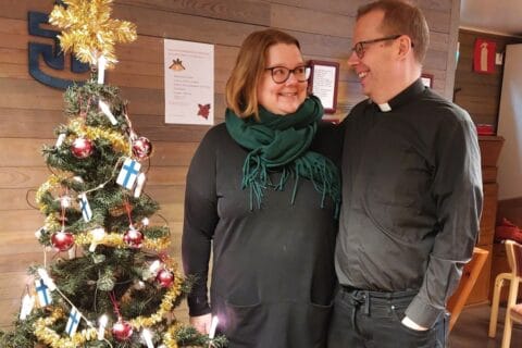 Toisiinsa katsovat, hymyilevät mustiin papinpaitoihin pukeutuneet nainen ja mies koristellun joulukuusen vieressä.