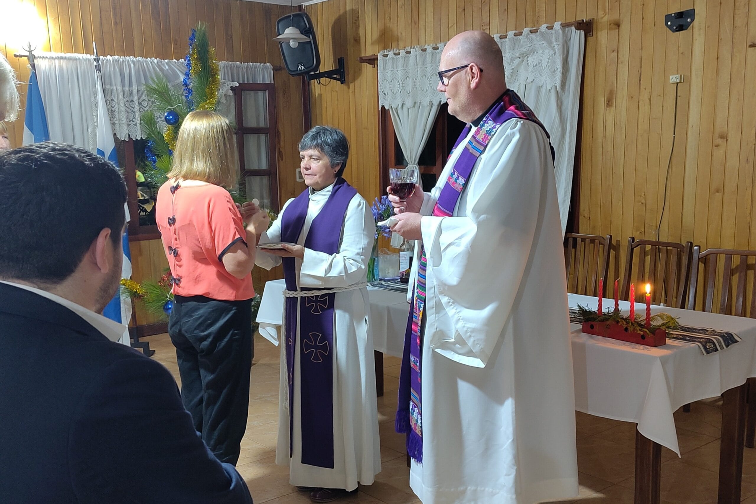 Valkoiseen papinalbaan pukeutuneet nainen ja mies jakavat ehtoollista vaaleahiuksiselle naiselle.