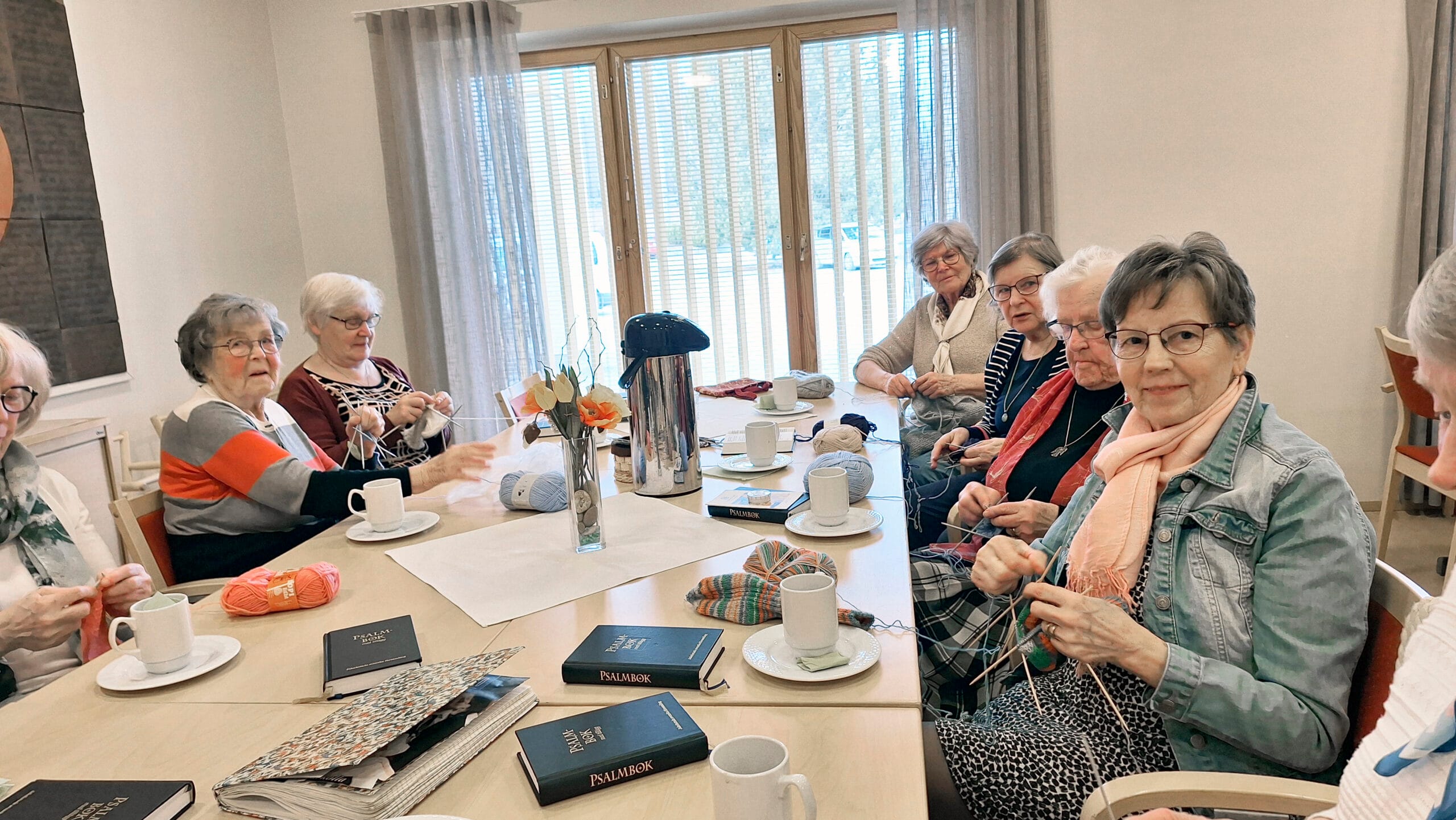 En grupp leende äldre kvinnor, varav några stickar, runt ett bord med kaffekoppar och psalmböcker.