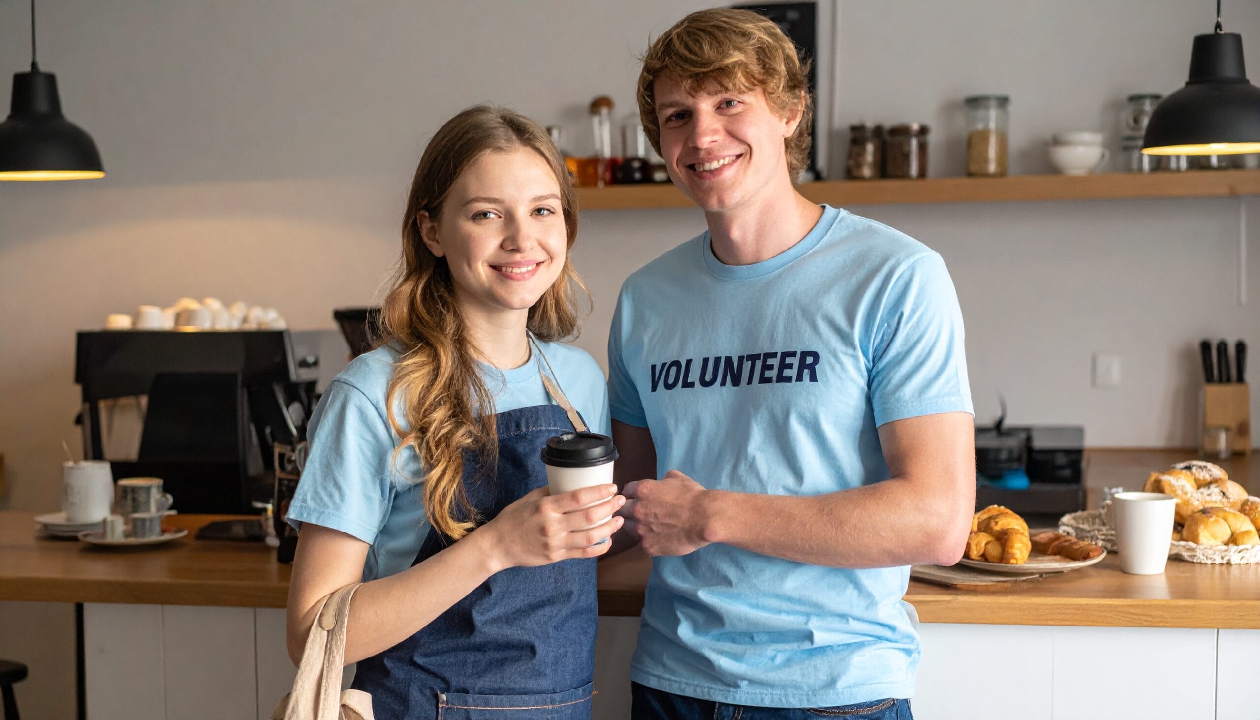Vaalea nuori mies yllään vaaleansininen paita, jossa lukee "volunteer" sekä hymyilevä nuori nainen seisovat kahvikuppi kädessään pöydän edessä, jossa on pullaa.