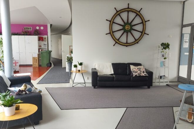 Sisätilakuva valkoisesta aulasta, jossa lattialla harmaat matot ja takana musta nahkasohva ja seinällä puinen iso laivan ruori.