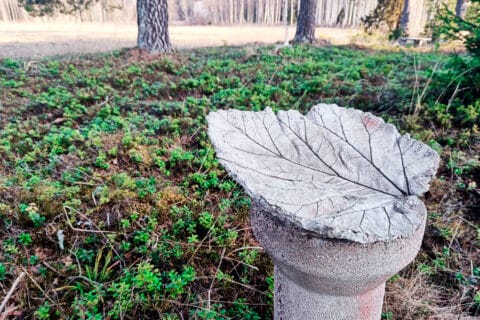 Betonista tehty harmaa isohko koivunlehden muotoinen kupera esine pyöreän noin metrin korkuisen betonijalustan päällä, taustalla alkukevään kunttakerrosta ja puita.