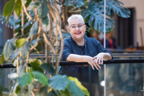 Hymyilevä, vaaleatukkainen ainen nojaa kaiteeseen viherkasveja ympärillään.