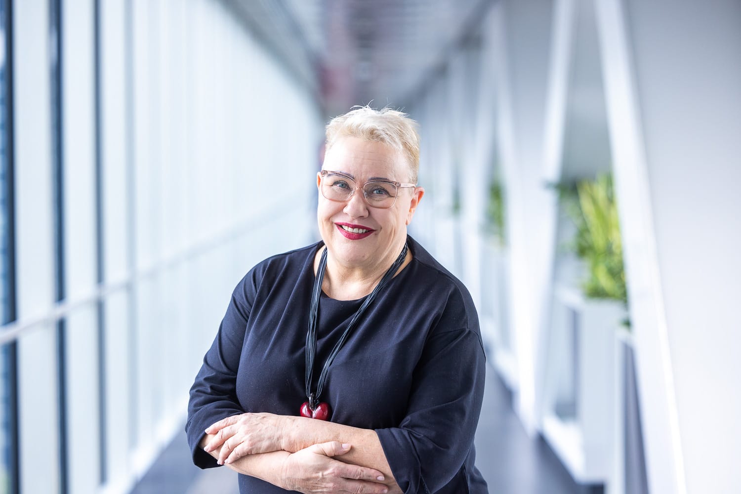 Vaaleatukkainen, silmälasipäinen hymyilevä nainen valoisassa lasikäytävässä.