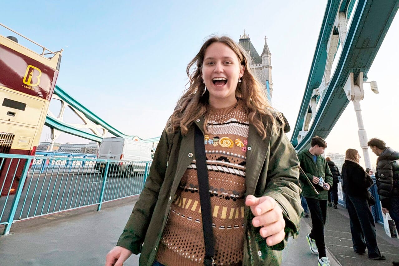 Nuori pitkähiuksinen hymyilevä nainen kävelee sillalla olallaan ja käsisään kasseja.