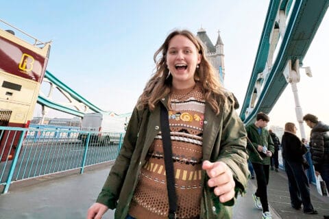 Nuori pitkähiuksinen hymyilevä nainen kävelee sillalla olallaan ja käsisään kasseja.