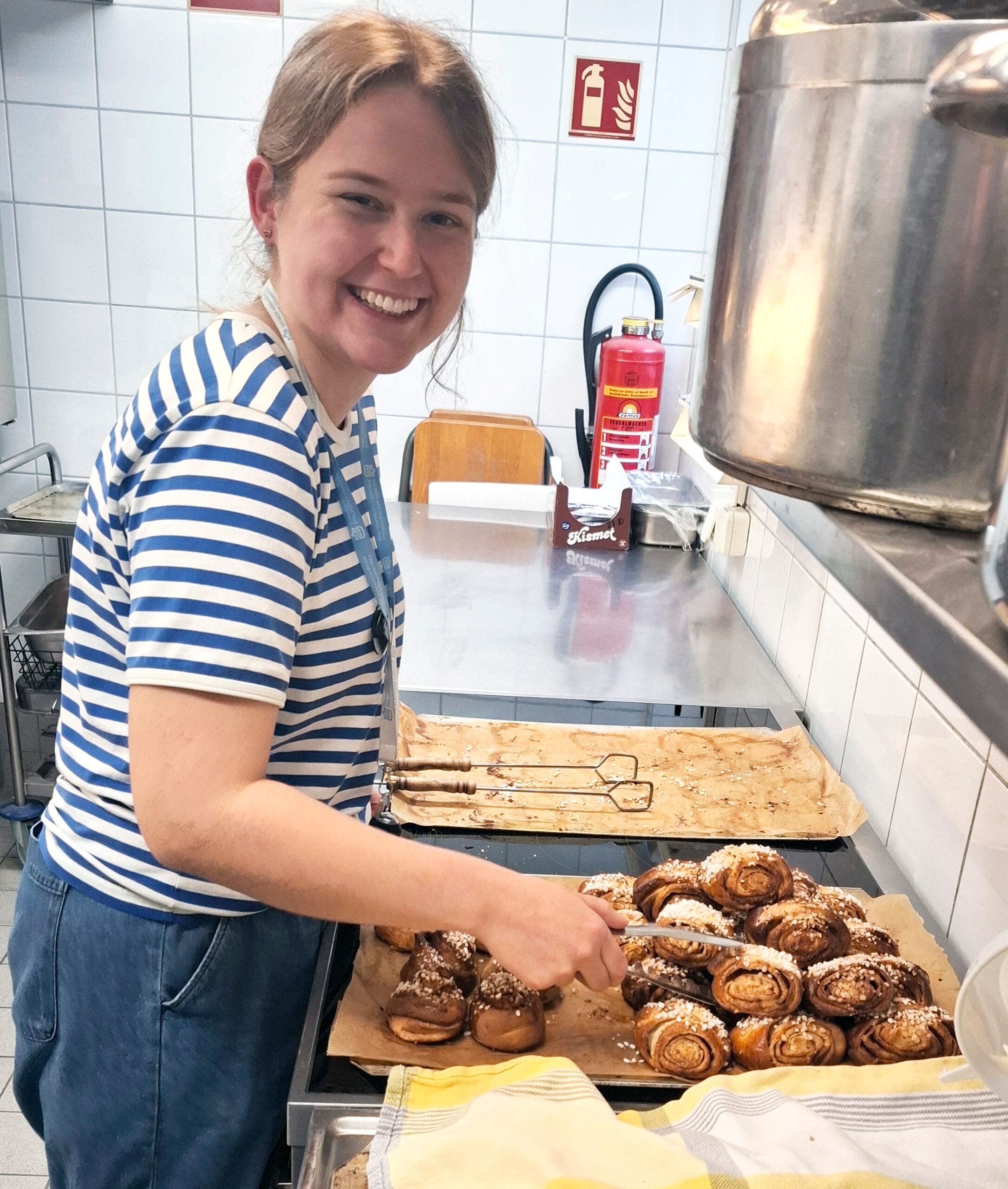 Hymyilevä, vaaleatukkainen nuori sinivalkoisessa raitapaidassa korvapuustipellin äärellä keittiötilassa. 