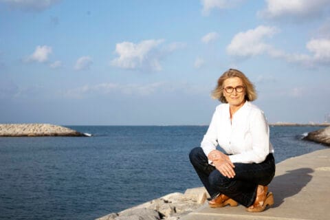 Vaaleahiuksinen nainen valkoisessa paidassa ja mustissa housuissa kyykyssä menrenrannan kalliolla.