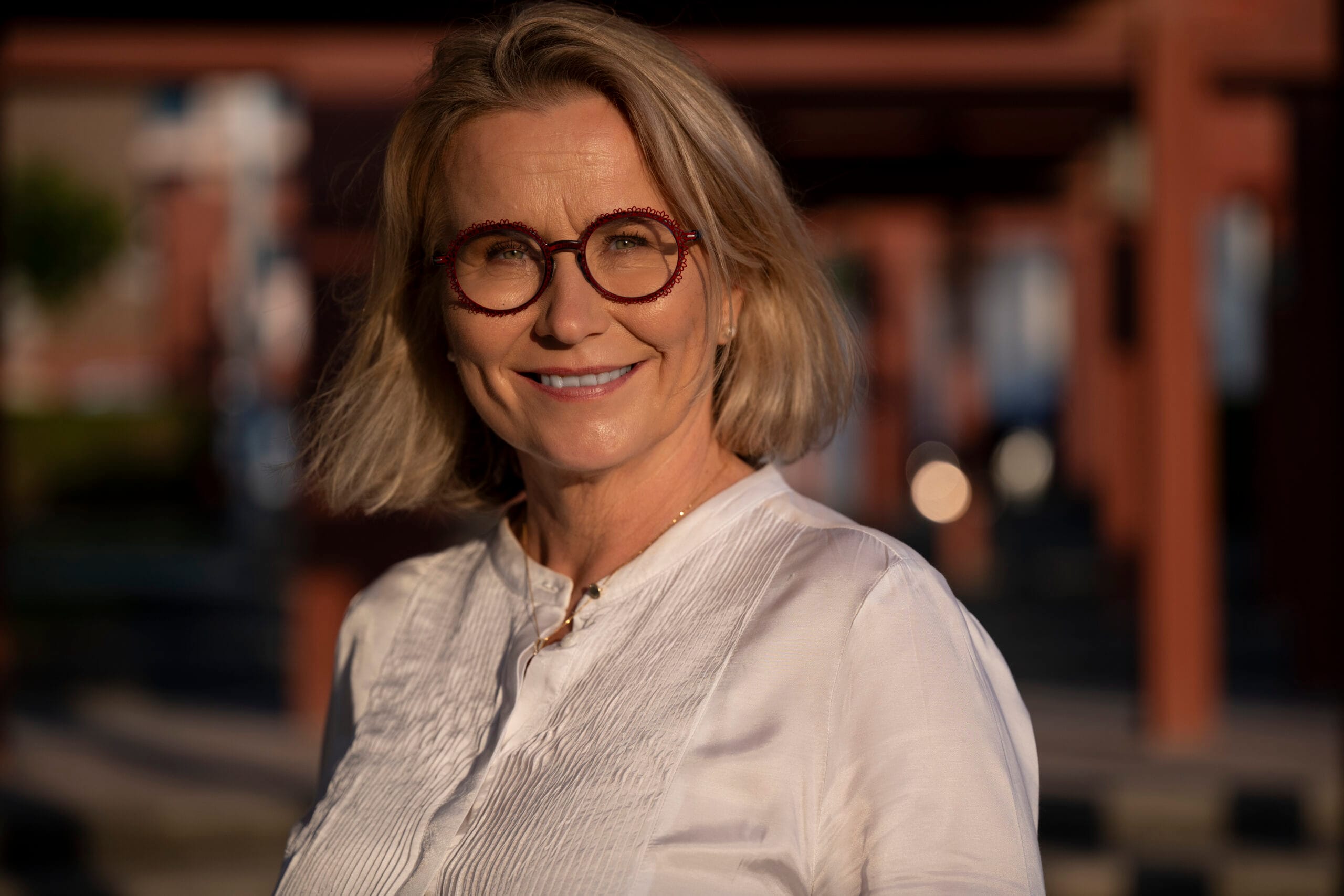 Hymyilevä, silmälasipäinen vaaleatukkainen nainen yllään valkoinen paita.