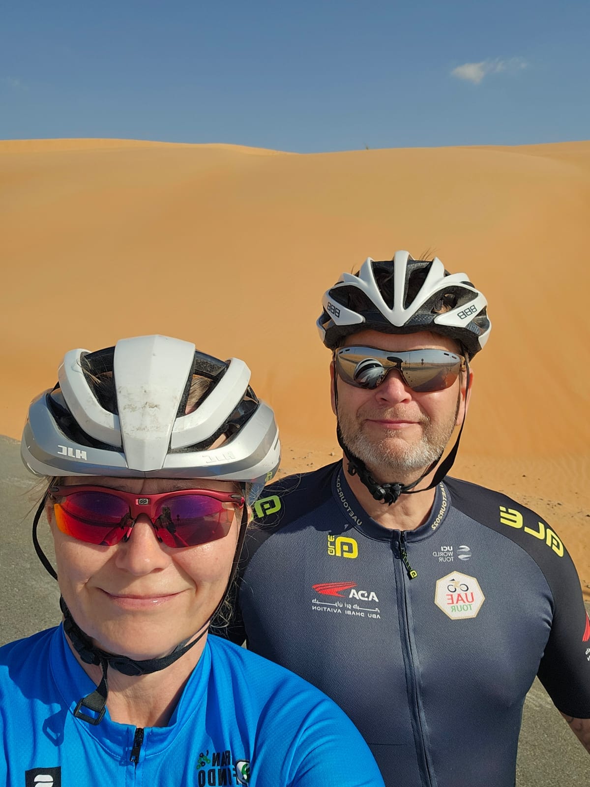 Hymyilevät nainen ja mies pyöräilykypärä päässään taustalla aavikon hiekkaa.