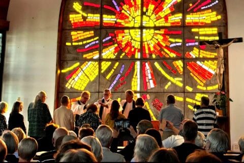 Takana iso, aurinkoa kuvaava keltapunainen lasi-ikkuna, jonka edessä kolme valkoiseen papin albaan pukeutunutta miestä jakaa ehtoollista joukolle ihmisiä.